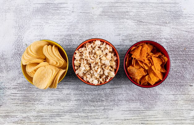 Top view of chips and popcorn in bowls on white wooden  horizontal