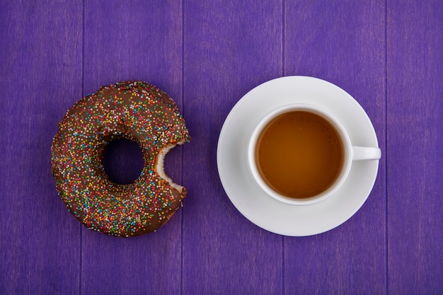 Vista dall'alto della ciambella morsicata al cioccolato con una tazza di tè su una superficie viola brillante