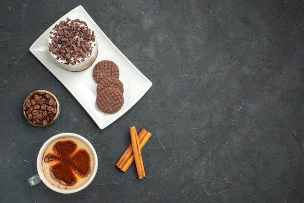 Top view chocolate cake and biscuits on white rectangular plate cup of coffee cinnamon sticks bowl with coffee seeds on dark isolated background