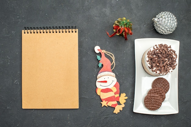 Top view chocolate cake and biscuits on white rectangular plate xmas tree toys a notebook on dark isolated background