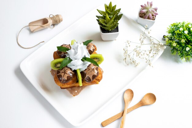 A top view chocolate cake designed with custard sliced kiwis inside white plate along with decoration plants on the white background biscuit cookie sweet