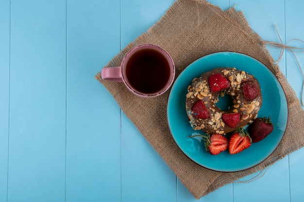 Top view of chocolate donut with strawberries on a blue plate with a cup of tea on a beige napkin on a blue surface