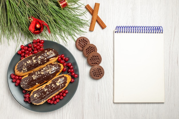 Вид сверху шоколадные эклеры и смородина на серой тарелке, печенье, скрещенные с корицей и сосновыми листьями с рождественскими игрушками и записной книжкой на белом деревянном столе