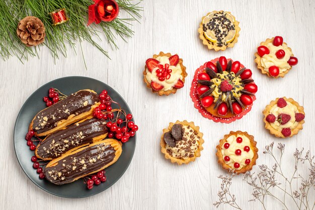 Top view chocolate eclairs and currants on the grey plate berry cake rounded with tarts and pine tree leaves with christmas toys on the white wooden ground