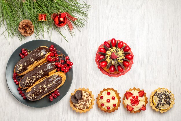 上面図チョコレートエクレアとスグリの灰色のプレートの下部にあるベリーケーキのタルトと松の木の葉とコピースペースのある白い木の地面にクリスマスのおもちゃ