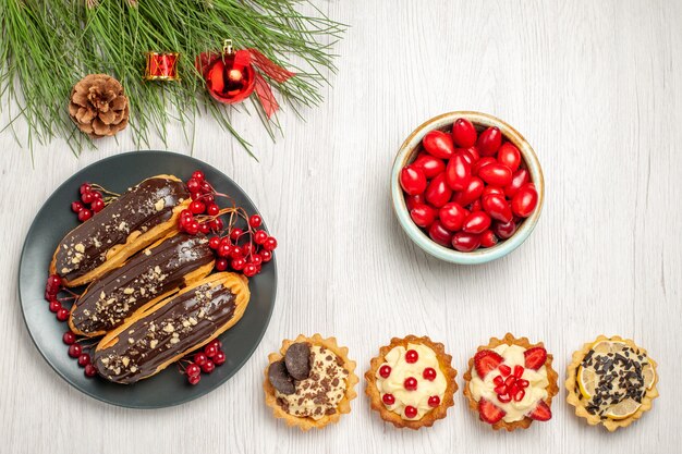 Top view chocolate eclairs and currants on the grey plate a bowl of cornel tarts and pine tree leaves with christmas toys on the white wooden ground with copy space