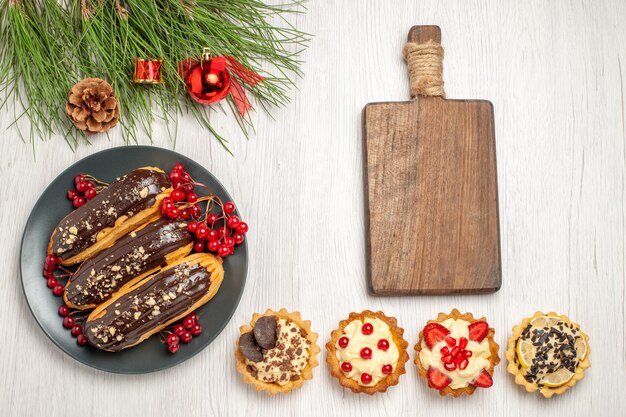 회색 접시에 상위 뷰 초콜릿 eclairs 및 건포도 도마 타르트와 소나무 흰색 나무 바닥에 크리스마스 장난감 나뭇잎