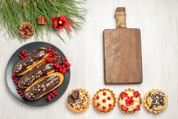Вид сверху шоколадные эклеры и смородина на серой тарелке, разделочная доска, пироги и сосновые листья с рождественскими игрушками на белой деревянной земле