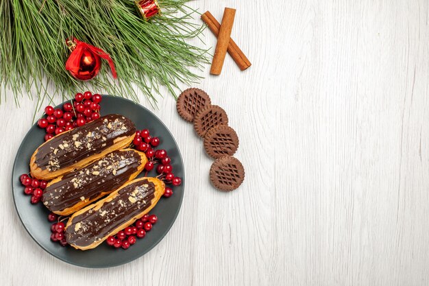 Вид сверху шоколадные эклеры и смородина на серой тарелке, печенье, скрещенные с корицей и сосновыми листьями с рождественскими игрушками на белом деревянном столе