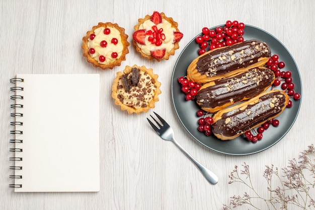 Top view chocolate eclairs and currants on the grey plate cookies a fork and a notebook on the white wooden table
