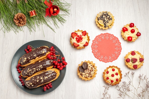 Top view chocolate eclairs and currants on the grey plate the red oval lace doily rounded with tarts and pine tree leaves with christmas toys on the white wooden ground