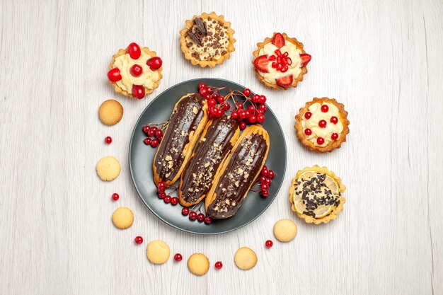 Top view chocolate eclairs and currants on the grey plate surrounded with tarts and cookies in the center of the white wooden table with copy space