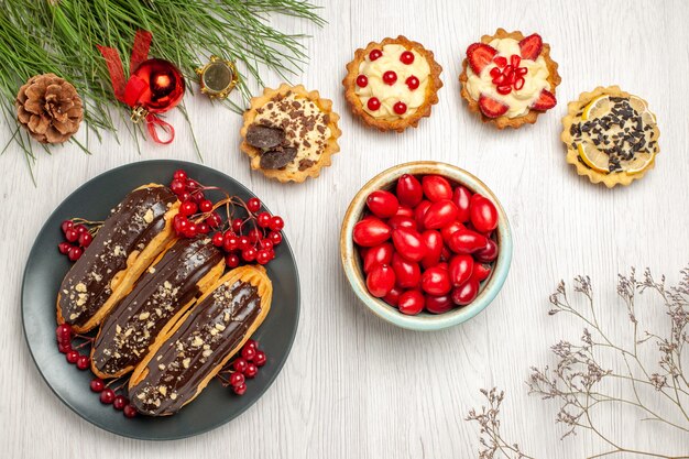 Top view chocolate eclairs and currants on the grey plate tarts a bowl of cornels and pine tree leaves with christmas toys on the