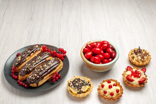 Top view chocolate eclairs and currants on the grey plate tarts and a bowl of cornels on the white wooden table with copy space