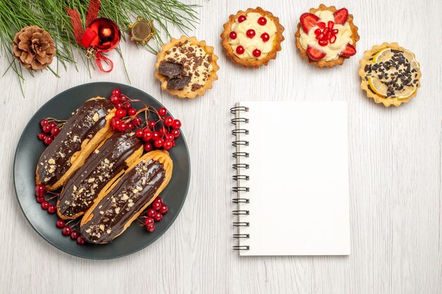 Top view chocolate eclairs and currants on the grey plate tarts a notebook and pine tree leaves with christmas toys on the white wooden table