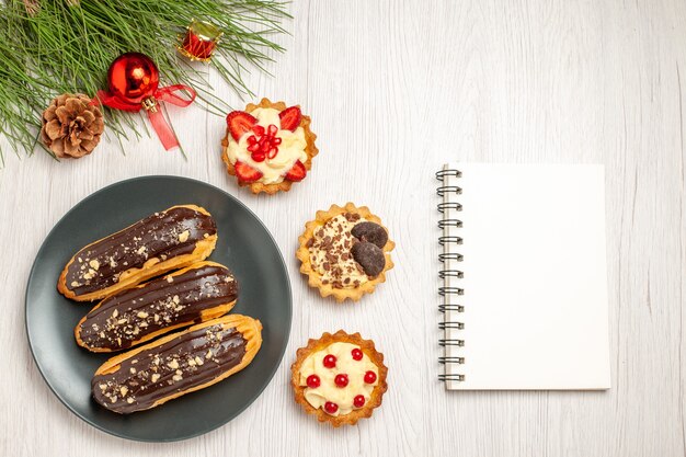 Top view chocolate eclairs on the grey plate tarts and the pine tree leaves with christmas toys at the left and a notebook at the right side of the white wooden ground