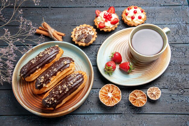 Top view chocolate eclairs on oval plate a cup o tea and strawberries on saucer tarts cinnamon and dried lemons on the dark wooden ground