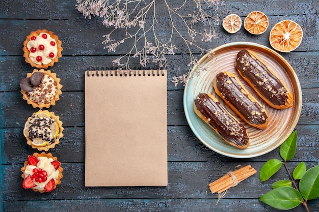 Top view chocolate eclairs on oval plate dried flower branch cinnamon dried oranges leaves a notebook and vertical row tarts on the dark wooden table