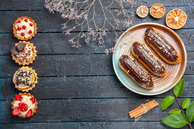 Top view chocolate eclairs on oval plate dried flower branch cinnamon dried oranges leaves and vertical row tarts on the dark wooden table with copy space
