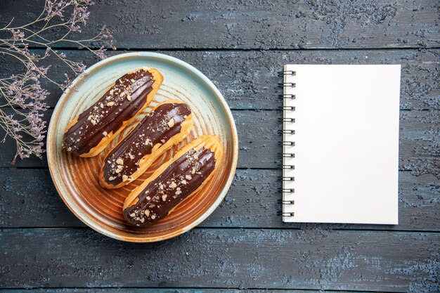 Top view chocolate eclairs on oval plate dried flower branch and a notebook on the dark wooden table