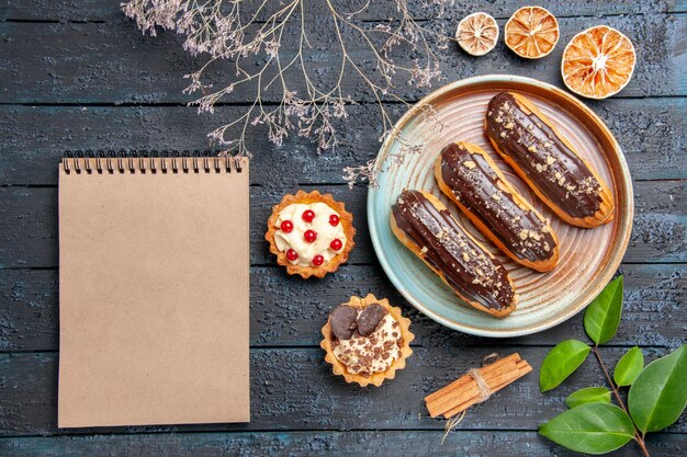 Top view chocolate eclairs on oval plate dried flower branch tarts cinnamon dried oranges leaves and a notebook on the dark wooden table