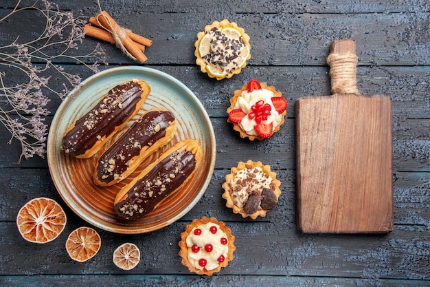 Top view chocolate eclairs on oval plate surrounded with dried lemons tarts and cinnamon and a chopping board on the dark wooden table