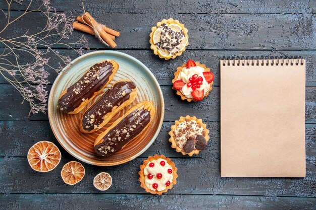 Top view chocolate eclairs on oval plate surrounded with dried lemons tarts and cinnamon and a notebook on the dark wooden table with copy space
