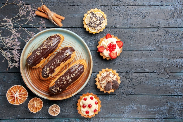 Top view chocolate eclairs on oval plate surrounded with dried lemons and tarts at the left side of the dark wooden table with copy space