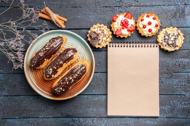 Top view chocolate eclairs on oval plate tarts cinnamon dried flower branch and a notebook on the dark wooden table