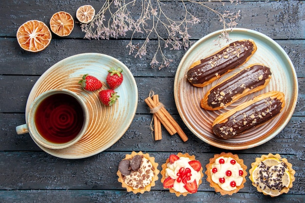 Top view chocolate eclairs on oval plate tarts cinnamon dried oranges and a cup of tea and strawberries on saucer on the dark wooden table