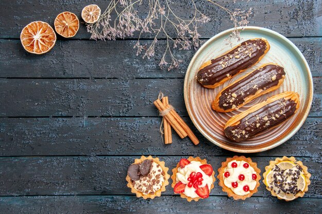 Top view chocolate eclairs on oval plate tarts cinnamon dried oranges on the dark wooden table with copy space