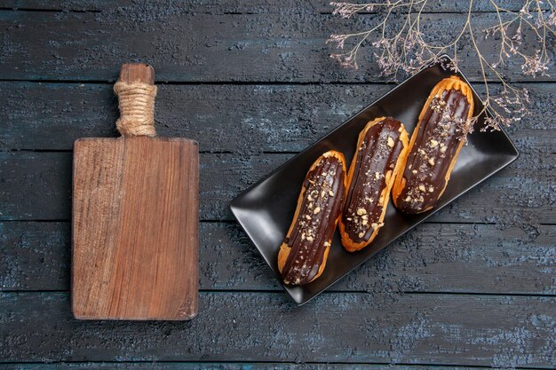 Top view chocolate eclairs on rectangle plate and a chopping board on the dark wooden table with free space