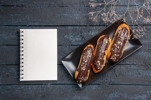 Top view chocolate eclairs on rectangle plate and a notebook on the dark wooden table with free space