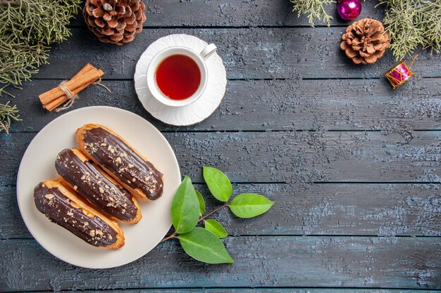 Top view chocolate eclairs on white oval plate cones christmas toys fir-tree leaves cinnamon and a cup of tea on dark wooden ground with copy space