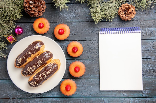 Вид сверху шоколадные эклеры на белой овальной тарелке еловые ветки рождественские игрушки вишневые кексы и тетрадь на темном деревянном столе