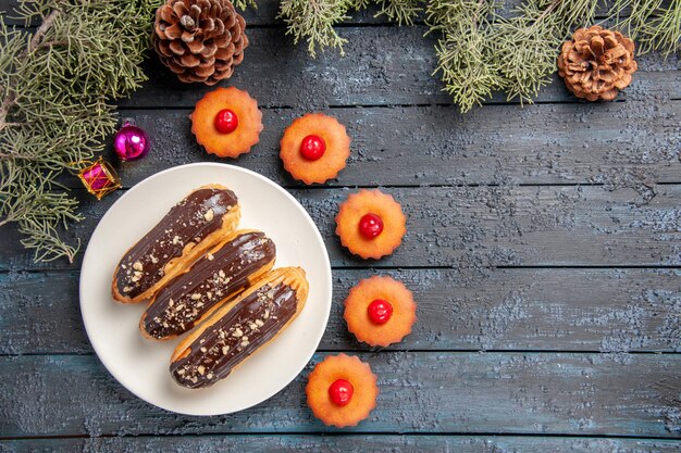 Top view chocolate eclairs on white oval plate fir-tree branches christmas toys and cupcakes on dark wooden table with copy space