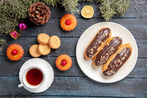 Vista dall'alto bignè al cioccolato su piastra ovale bianca rami di abete giocattoli di natale fetta di limone una tazza di biscotti da tè e cupcakes su fondo di legno scuro