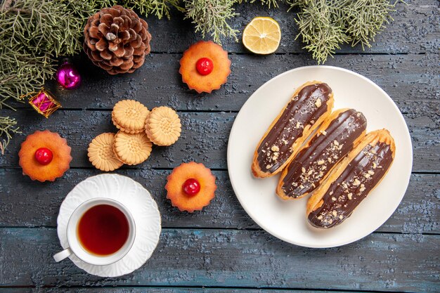 Top view chocolate eclairs on white oval plate fir-tree branches christmas toys slice of lemon a cup of tea biscuits and cupcakes on dark wooden ground