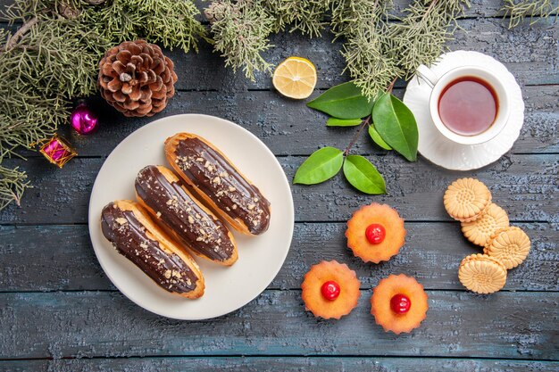Top view chocolate eclairs on white oval plate fir-tree branches christmas toys slice of lemon a cup of tea biscuits and cupcakes on dark wooden table