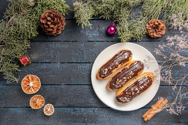 흰색 타원형 접시 전나무 나뭇 가지와 콘 크리스마스 장난감에 상위 뷰 초콜릿 eclairs 여유 공간이있는 어두운 나무 테이블에 오렌지 계피를 건조