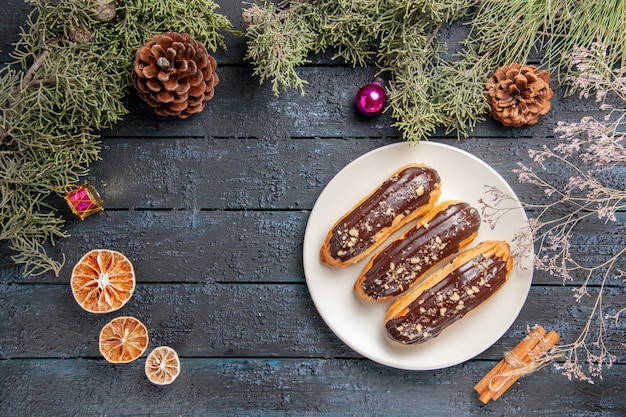 Вид сверху шоколадные эклеры на белой овальной тарелке еловые ветки и шишки рождественские игрушки сушеные апельсины корица на темном деревянном столе со свободным пространством