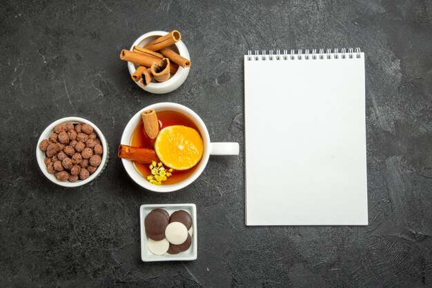 Top view chocolate hizelnuts white notebook and bowls of cinnabon sticks chocolate and hizelnuts next to the cup of tea with cinnabon and lemon on the left side of the table