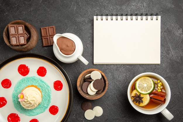 Top view chocolate on the table cupcake with cream and sauces next to the cup of herbal tea and white notebook bowls of chocolate and chocolate cream on the dark table