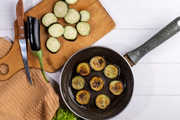 Top view chopped fried eggplant in a pan with chopped fresh eggplant on a board with a knife