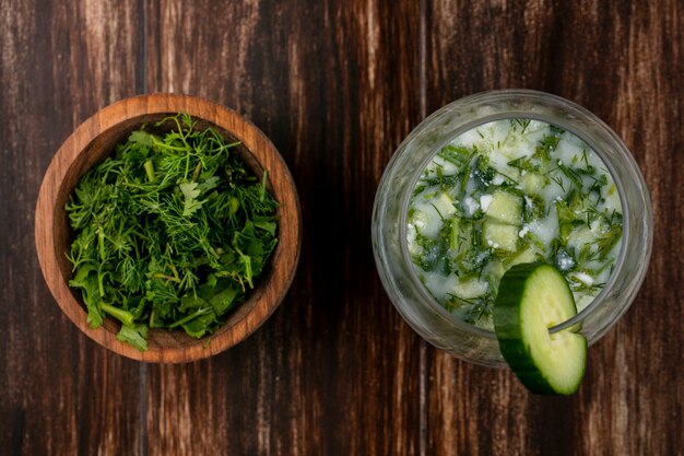 Top view of chopped greens with okroshka and cucumber on a wooden surface