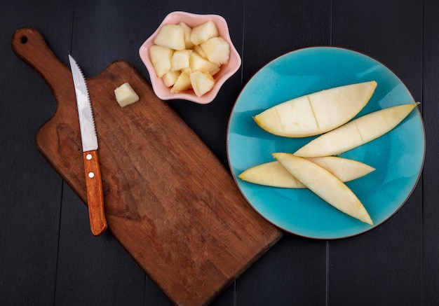 Вид сверху нарезанных ломтиков дыни на розовой миске с кожурой на синей тарелке с деревянной кухонной доской с ножом на черном
