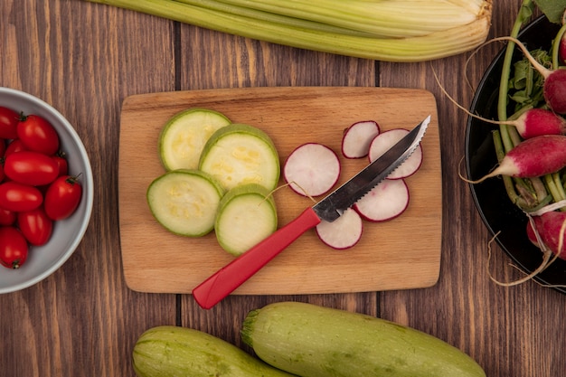Top view of chopped slices of zucchini and radish on a wooden kitchen board with knife with plum tomatoes on a bowl with radishes on a bowl with zucchini and celery isolated on a wooden background