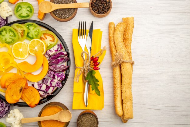 Top view chopped vegetables and fruits pumpkin bell peppers persimmon red cabbage green tomatoes on black plate various spices in bowls fork and knife on yellow napkin bread on white table