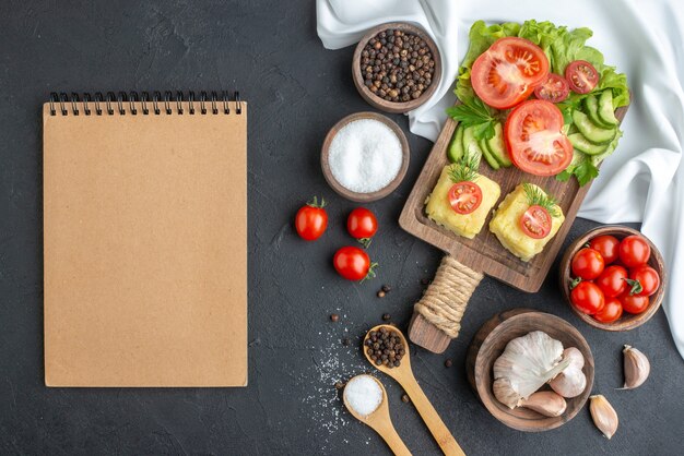 Top view of chopped and whole fresh vegetables on cutting board in bowls and spices notebook on white towel on black surface