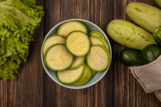 Top view of chopped zucchinis on a bowl with fresh vegetables such as cucumbers and zucchinis on a burlap bag with lettuce isolated on a wooden background